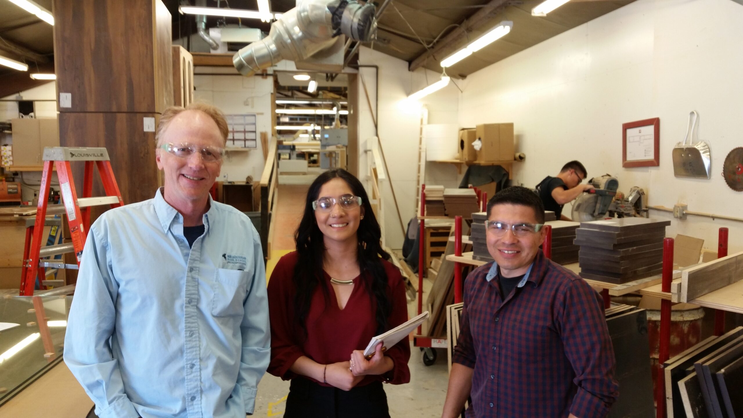 a student stands with her two adult mentors in a workshop, all three wear protective eye wear and hold clipboards