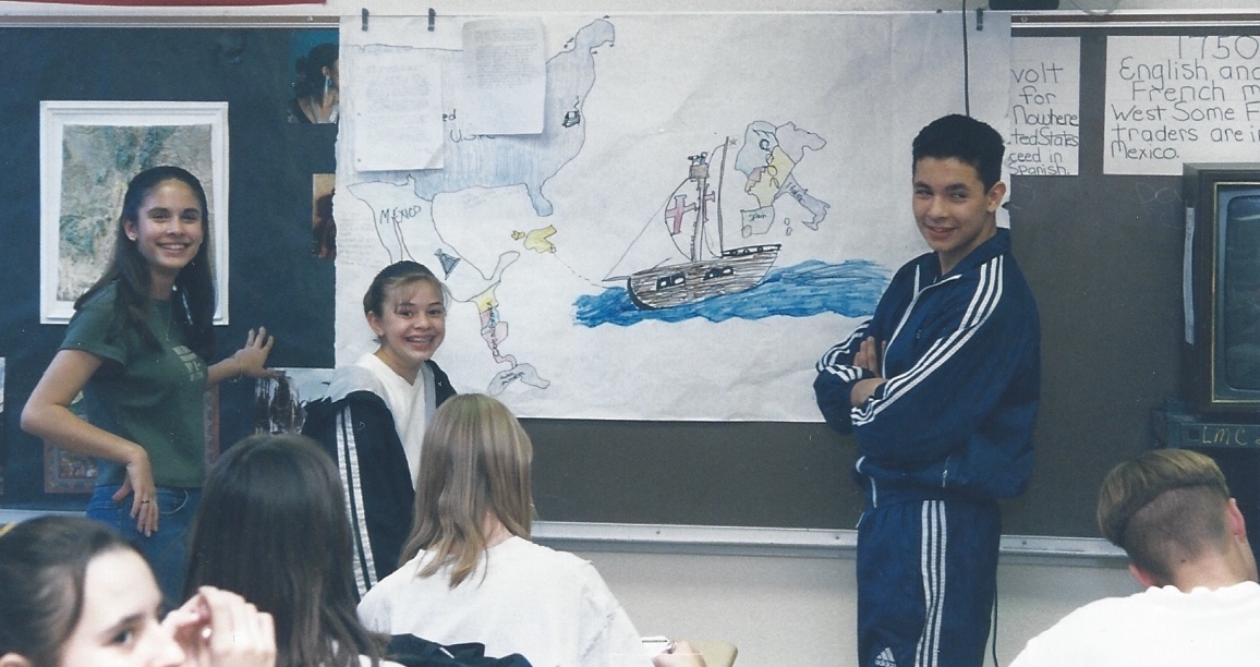 students stand by a white board smiling, presenting a history project to the class in the background