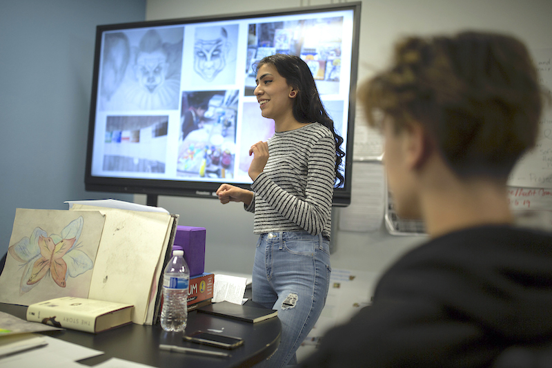 A female student in a striped sweater stands in front of a large presentation screen talking to a room full of teachers and mentors.
