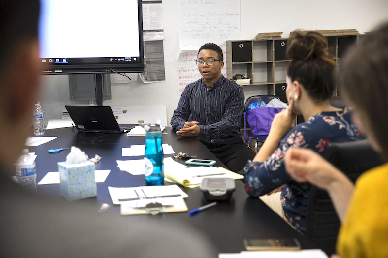 A male student in a formal collared shirt sits at a large table answering questions from his mentors and teachers.