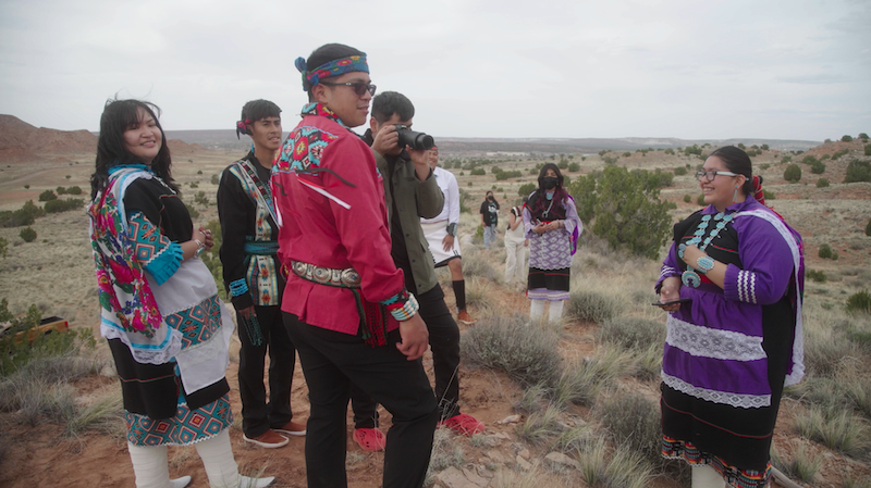 Zuni high school students stand in a group during a photography project for their senior capstone, they are wearing colorful traditional dress, with a desert background.