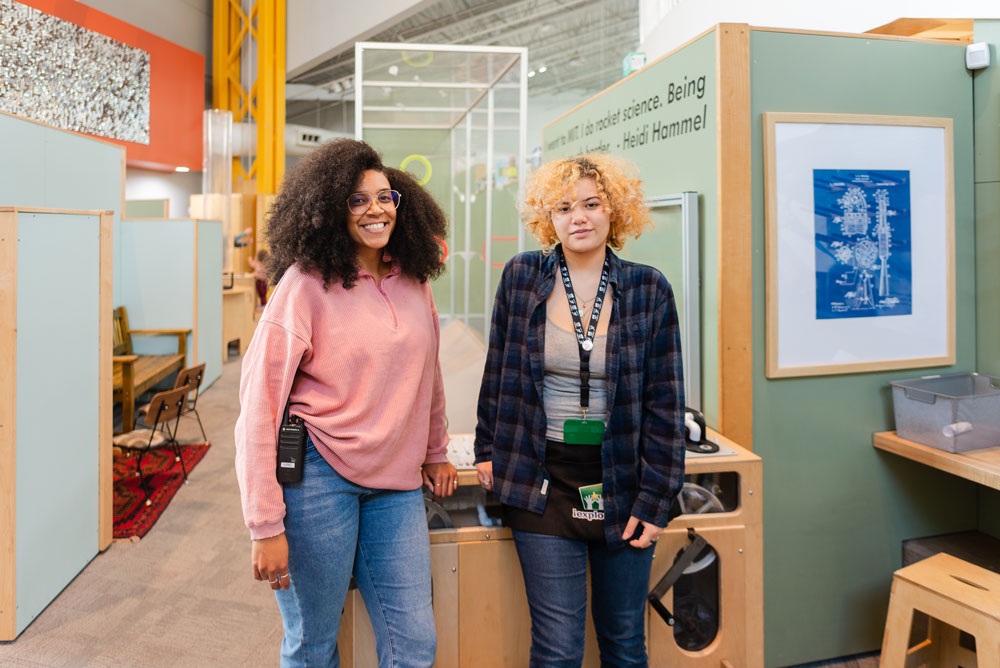 A student and mentor pose in front of an exhibit at Explora.