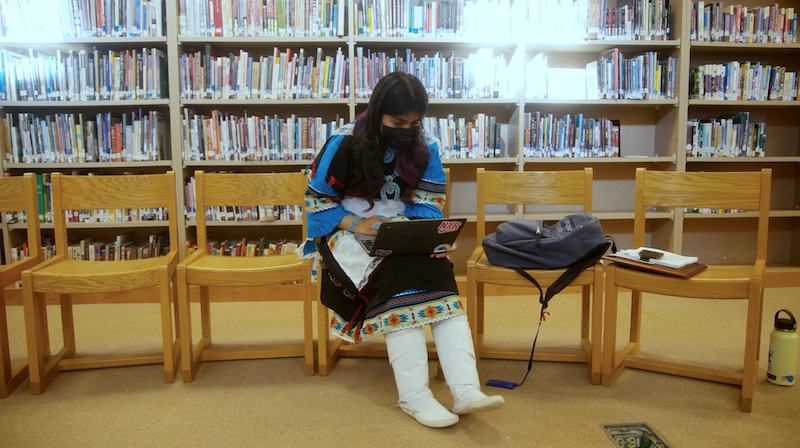 A student at Zuni High School sits in the library with her laptop wearing traditional ceremonial dress