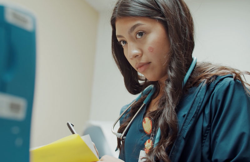 A high school student wears a stethoscope and writes notes on a clipboard at the doctor's office where she is an intern.