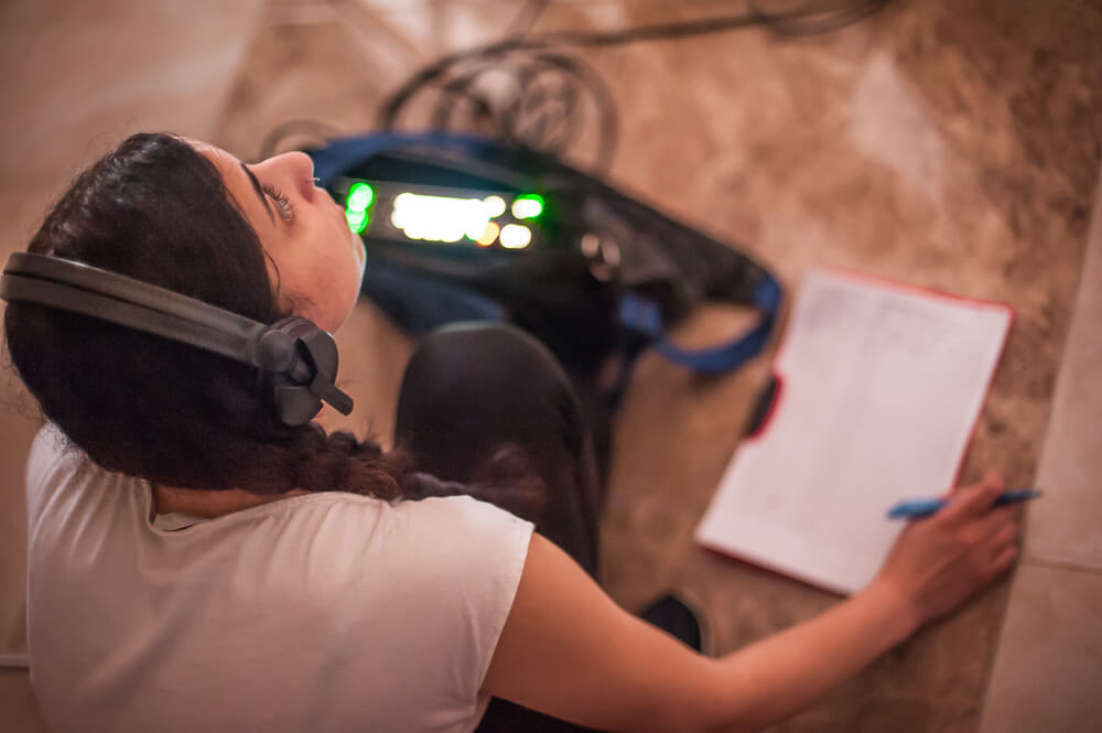 A student recording a Podcast, with headphones on, speaking into a microphone, referencing a notepad with writing on it.