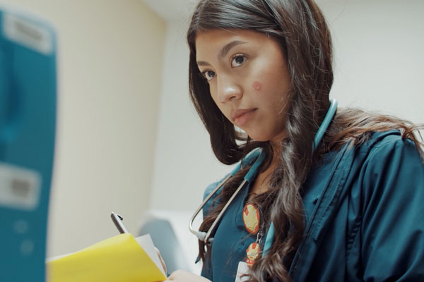 A high school student wears a stethoscope and writes notes on a clipboard at the doctor's office where she is an intern.
