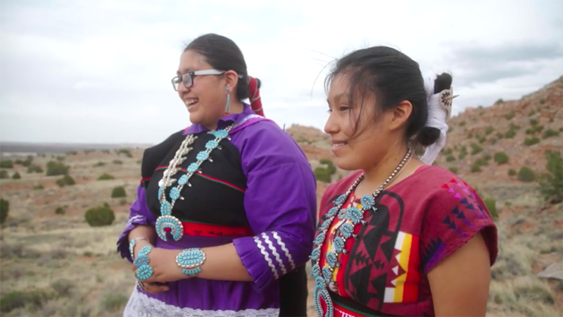 Two smiling indigenous high school students dressed in tribal regalia standing in a New Mexico desert landscape.
