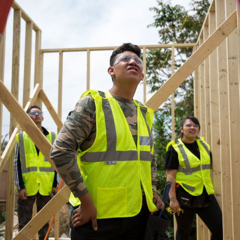 three students wearing construction vests and standing inside of a wooden structure, looking up.