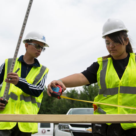 Two students wearing construction vests and helmets, working outside with measuring tools.