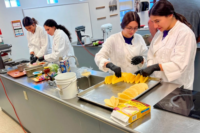 A group of four students wearing chef jackets and gloves preparing food