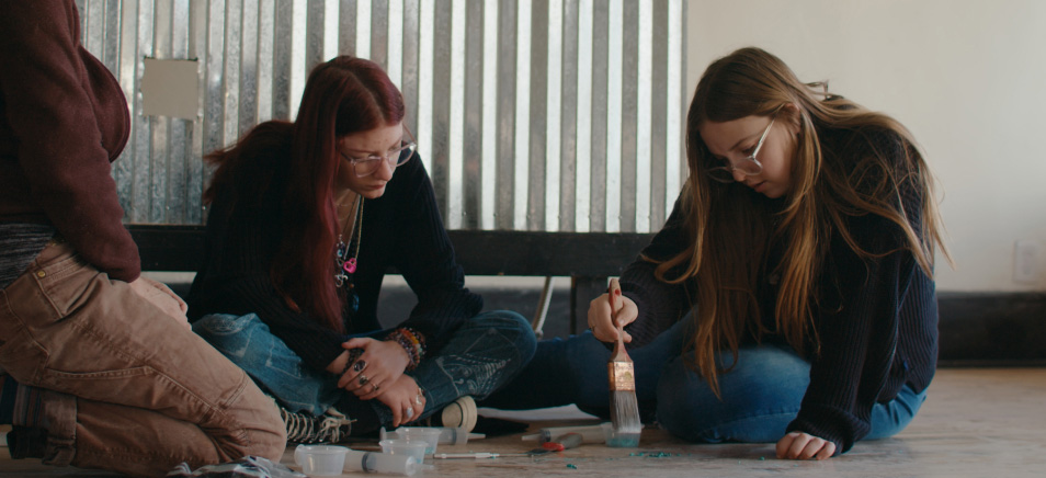 Two students sitting down on the floor, and one student using a paint brush