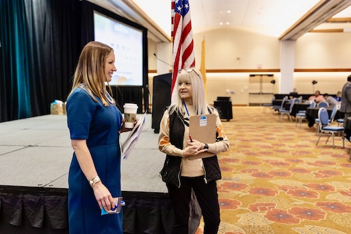 Two women speaking to each other beside a stage at a conference.